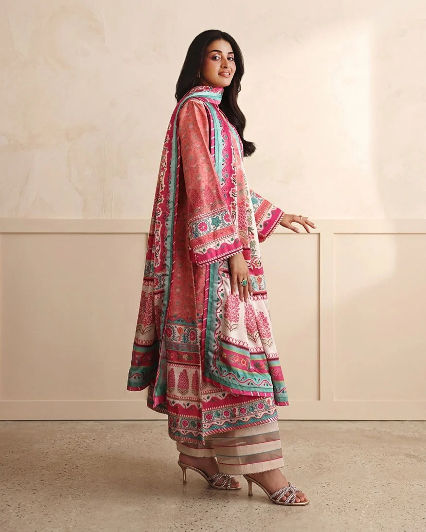 A woman models a traditional three-piece salwar kameez suit set in a warm beige studio setting. The set features a pink printed kameez jacket with teal and white borders, matching striped trousers, and a coordinating scarf.