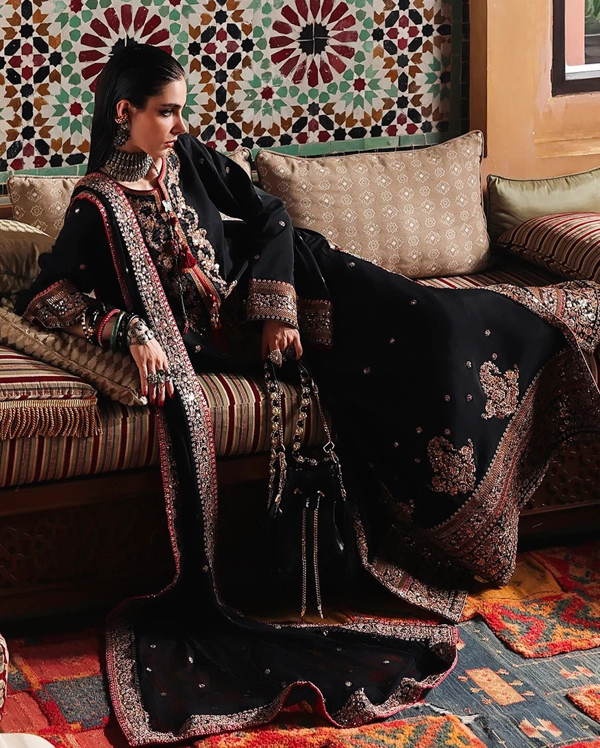 A woman seated on a patterned sofa wearing an ornate black traditional outfit with heavily embroidered borders in metallic and threadwork, set against a richly decorated interior with patterned cushions and rugs.