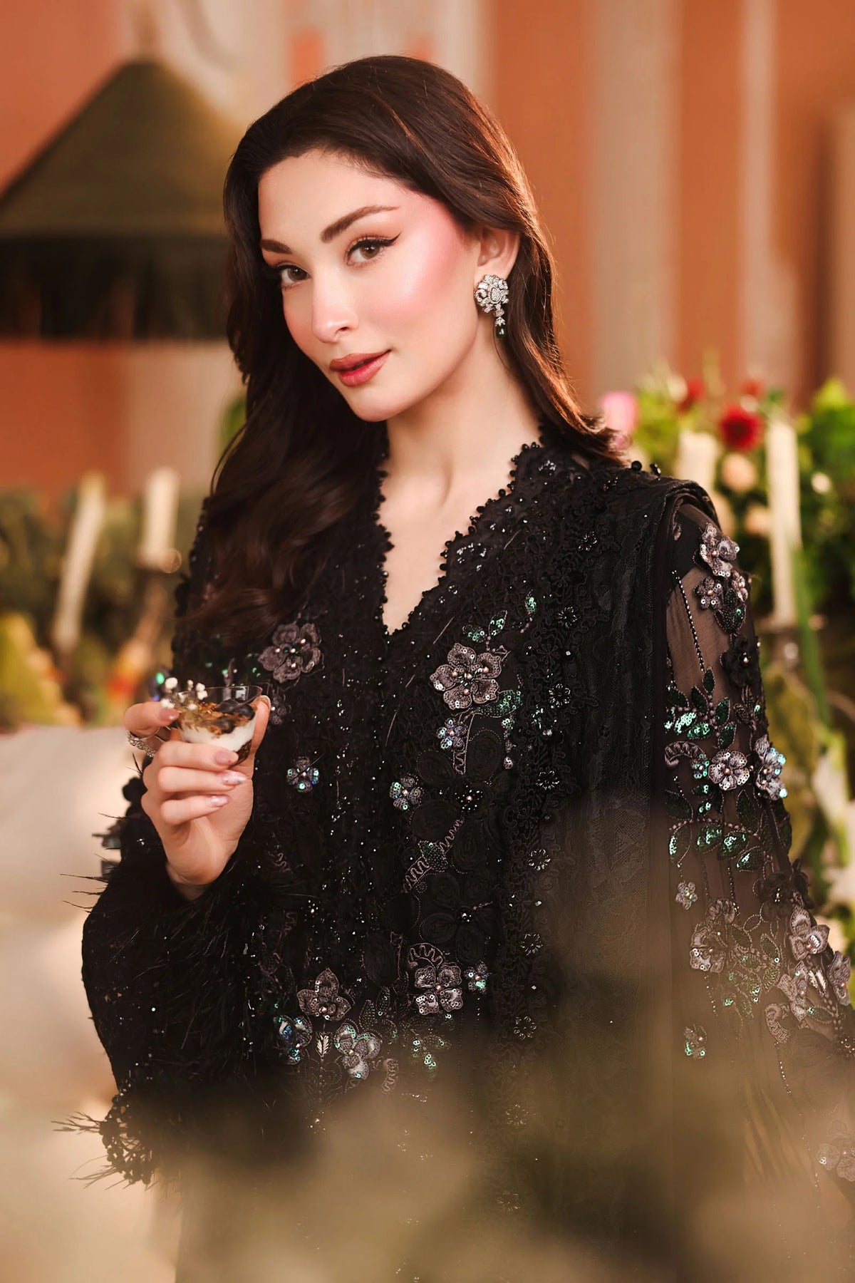 A woman wearing a black embellished lace dress with floral beadwork and sheer sleeves, holding a small glass in a softly lit indoor setting.