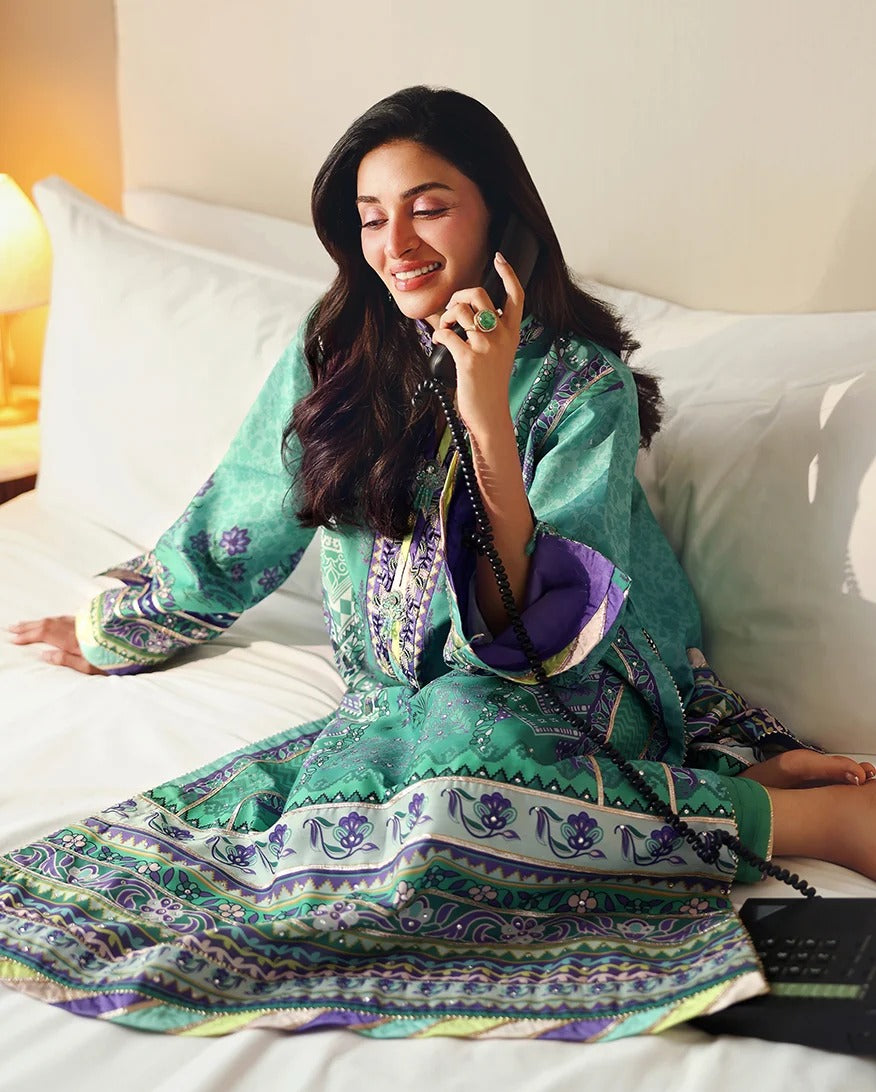 A woman sitting on a bed wearing a green and purple patterned traditional outfit, holding a phone to her ear.