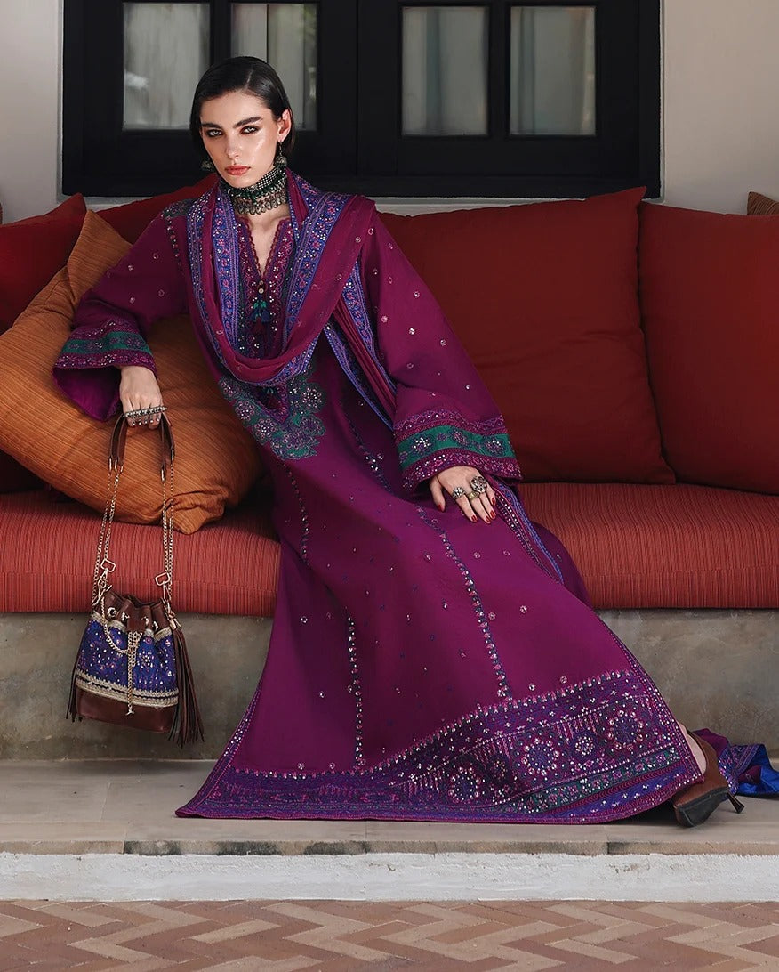 A woman models a deep purple embroidered traditional outfit with a matching dupatta, seated on a rust-red sofa. The outfit features intricate silver and blue embroidery along the borders and sleeves.