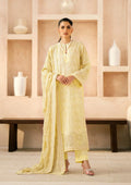 A woman modeling a light yellow embroidered traditional outfit with a matching dupatta, standing in front of a beige wall with built-in shelves and white vases.