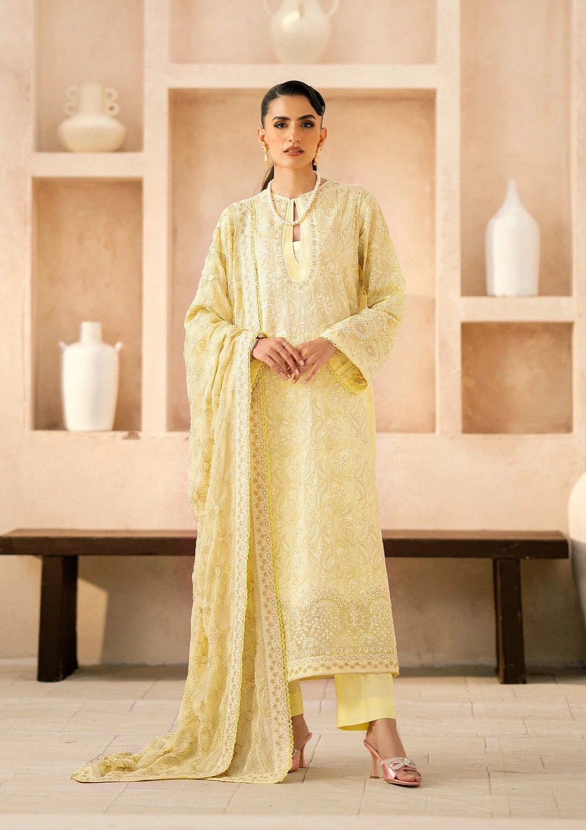 A woman modeling a light yellow embroidered traditional outfit with a matching dupatta, standing in front of a beige wall with built-in shelves and white vases.