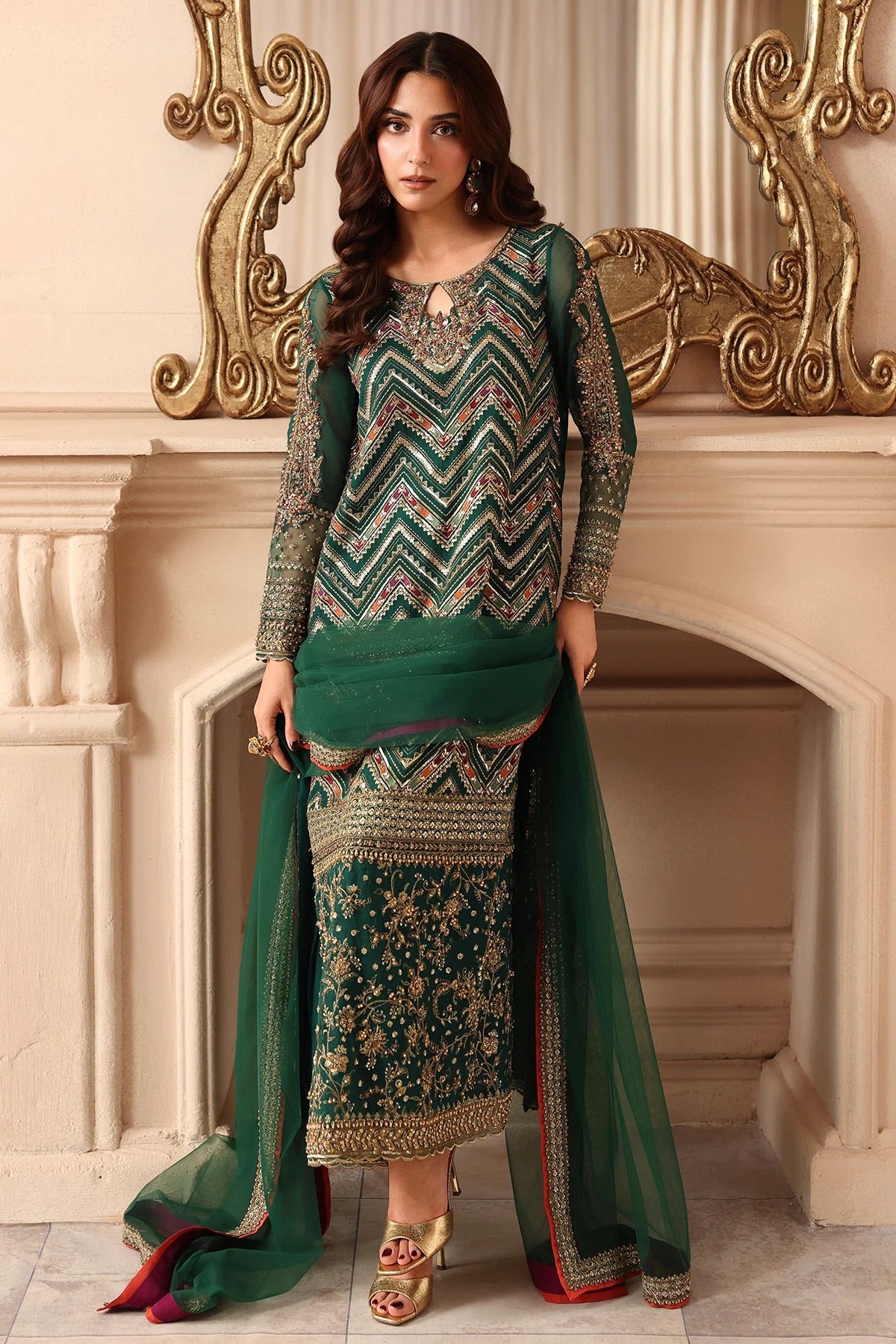 A woman models a traditional Indian salwar kameez suit in emerald green with intricate gold and beige embroidery. The set includes a long-sleeved kameez top with a chevron pattern, a matching flared kurta, and a sheer dupatta draped over her arm, set against an elegant beige wall with a gold decorative mirror.