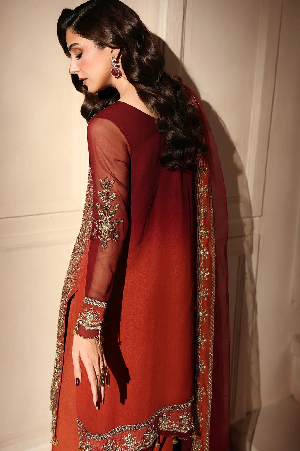 A woman wearing a deep red traditional South Asian outfit with sheer sleeves, ornate gold embroidery, and a matching dupatta, posed against a beige paneled wall.