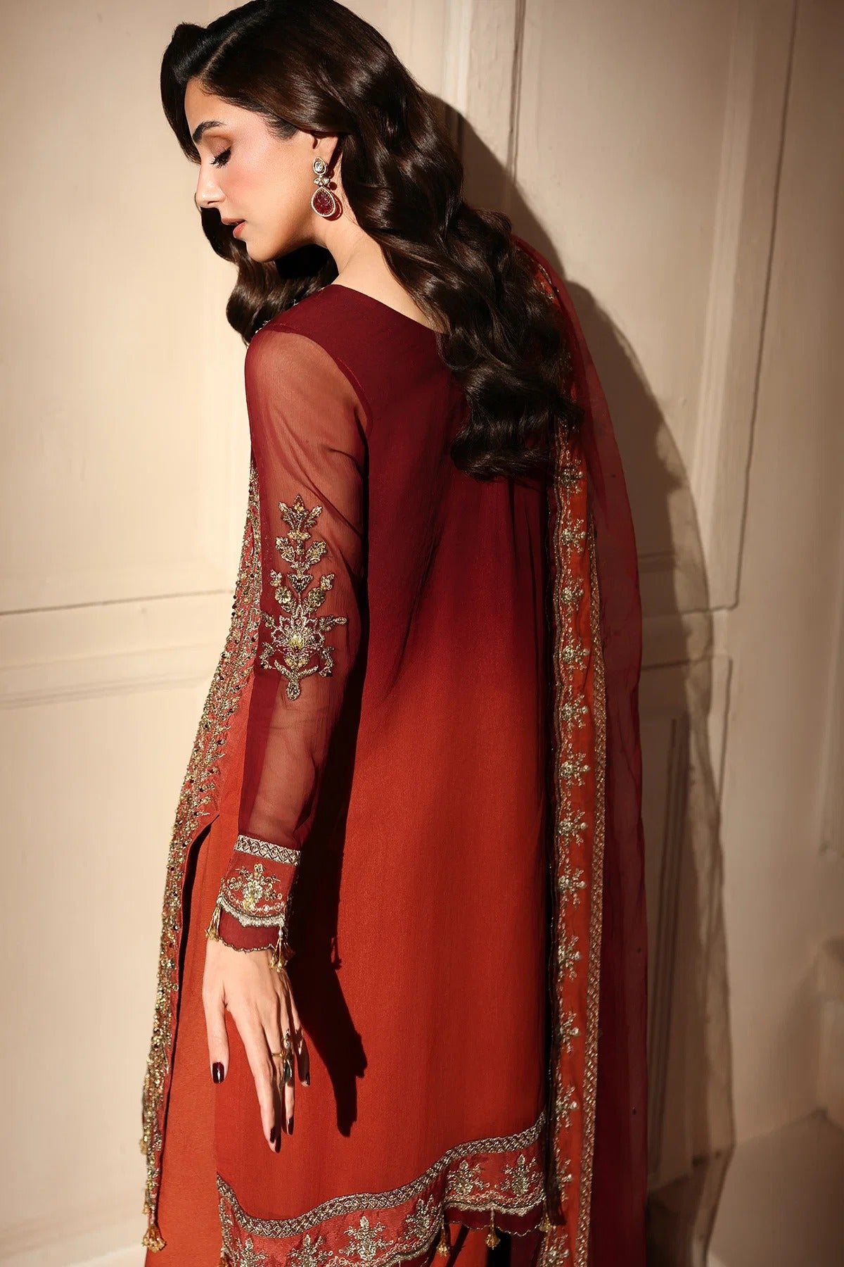 A woman wearing a deep red traditional South Asian outfit with sheer sleeves, ornate gold embroidery, and a matching dupatta, posed against a beige paneled wall.