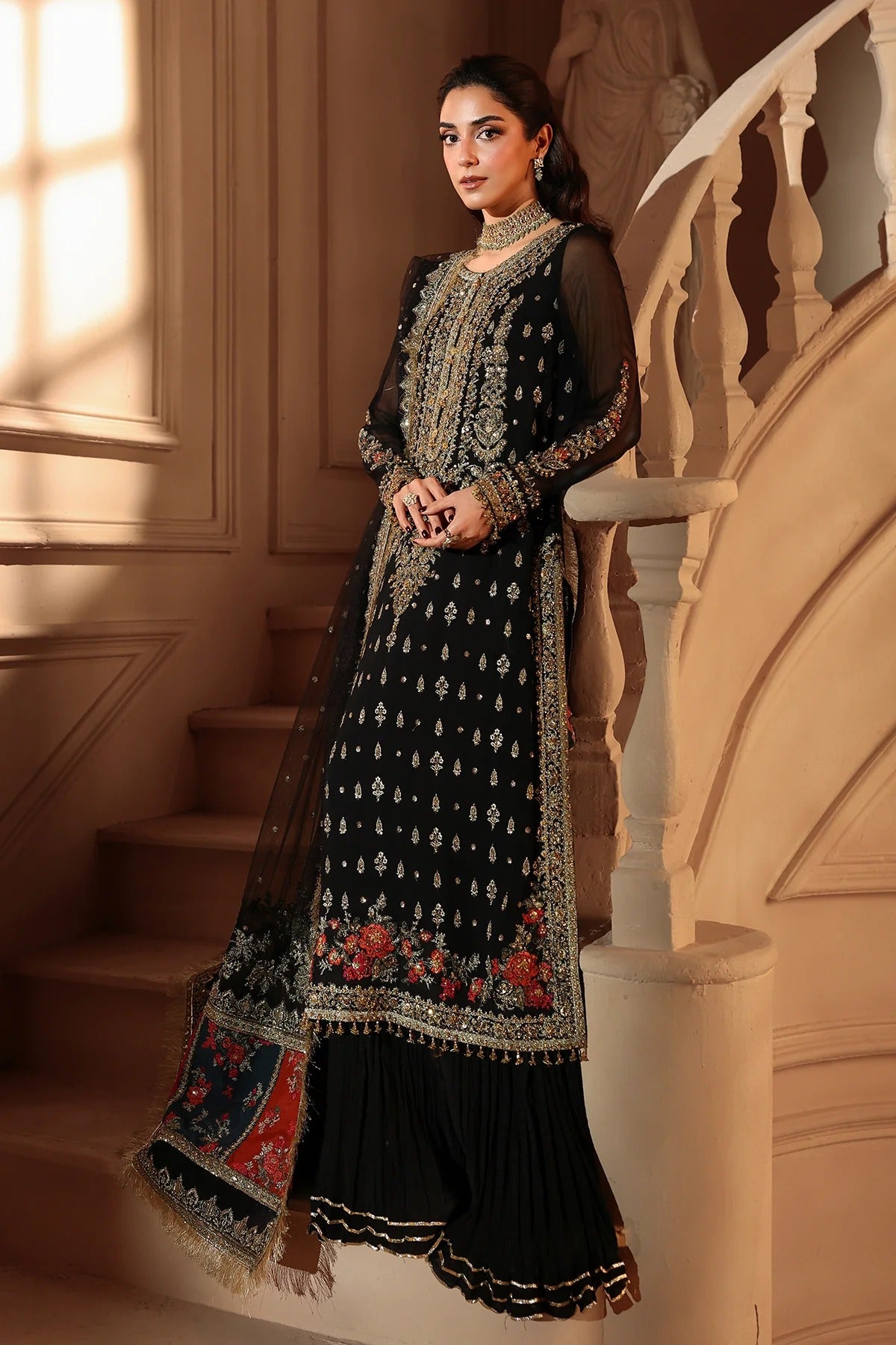 A woman models a black traditional South Asian outfit with intricate gold embroidery and red floral accents, standing on a beige staircase.