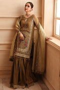 A woman models an elegant olive green and gold embroidered traditional outfit with a matching sheer dupatta, set against a warm beige wall next to a window.