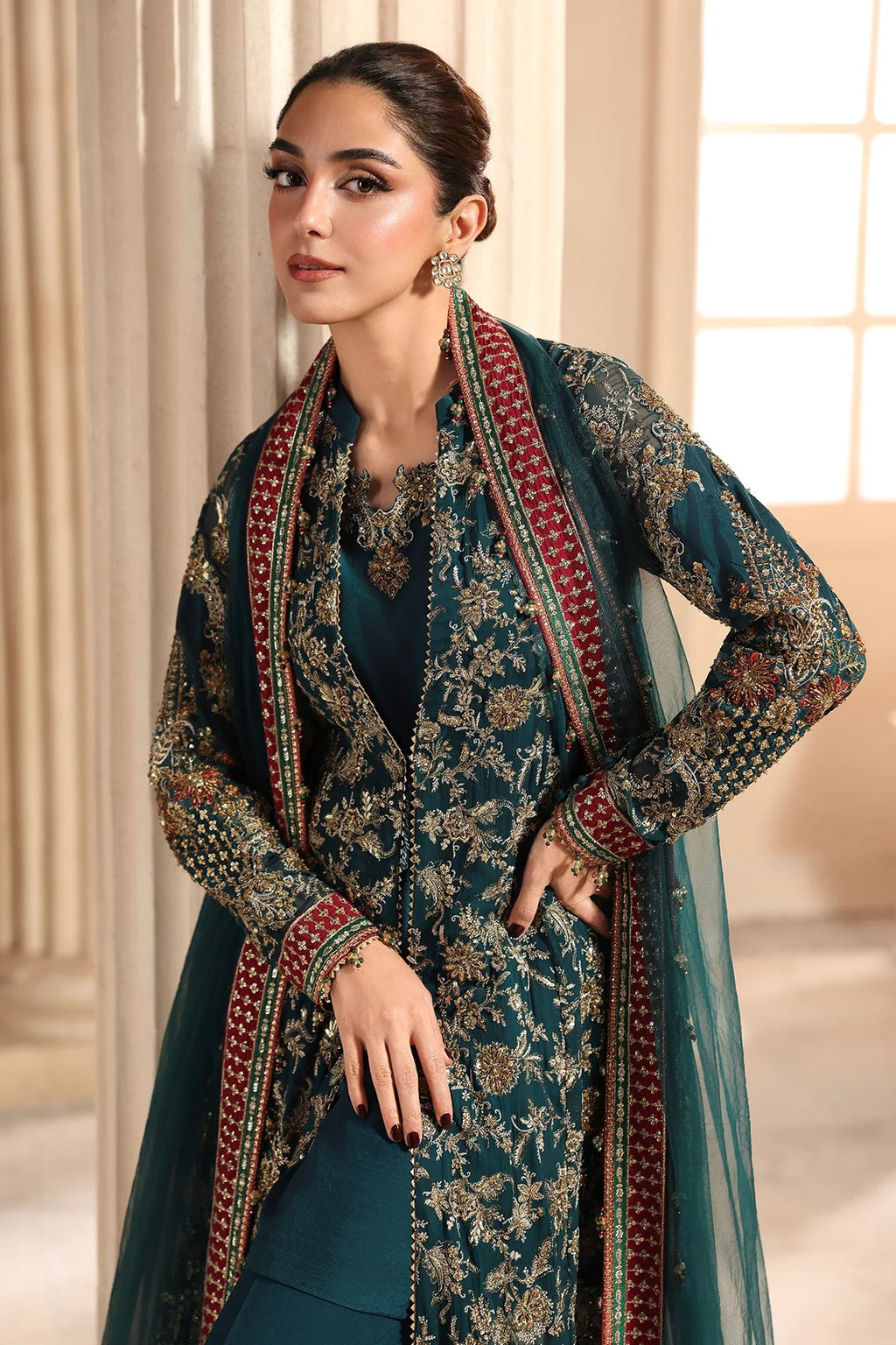 A woman modeling a dark teal green ethnic suit with intricate gold and red floral embroidery, featuring a matching sheer dupatta draped over her shoulders.