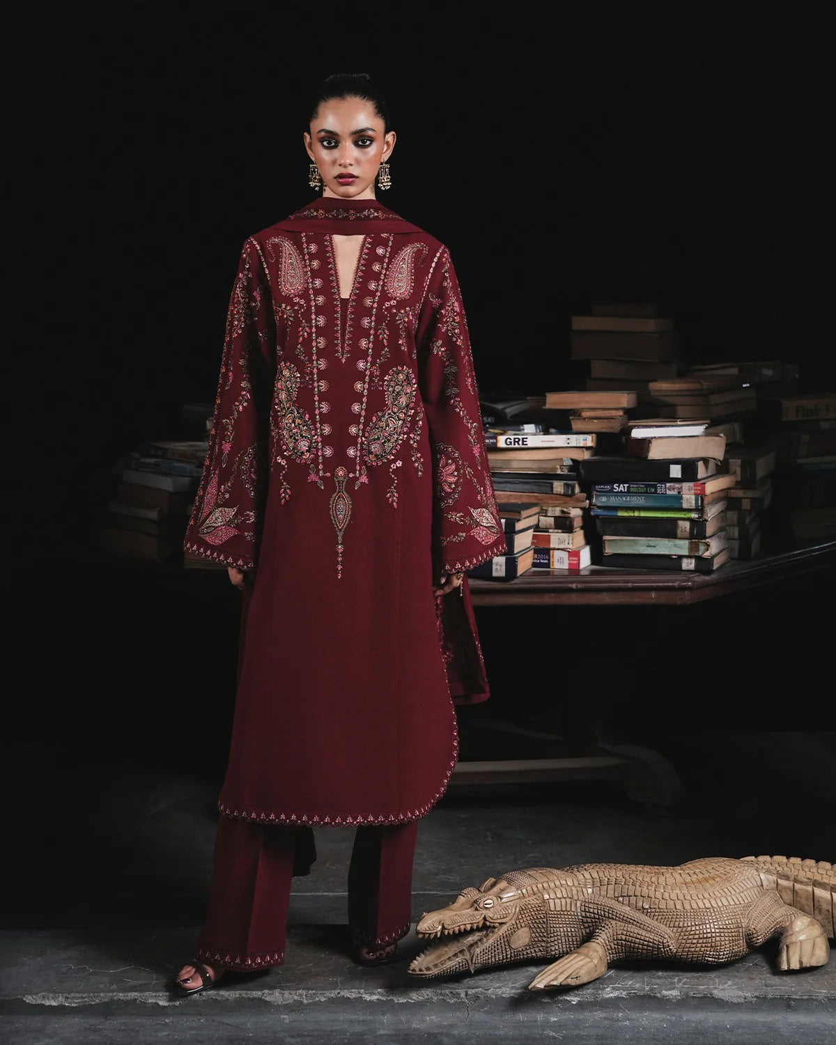 A model stands in a dark studio setting wearing a deep maroon embroidered kurta and matching trousers. The outfit features intricate gold and silver embroidery on the front and sleeves.