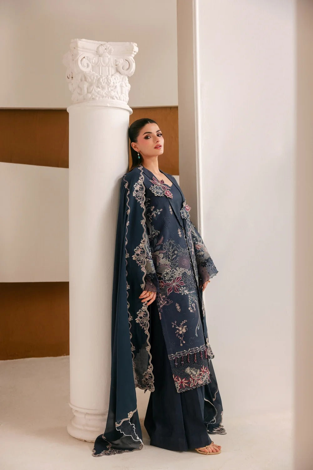 A woman models a dark navy blue traditional outfit with intricate floral embroidery and lace details, standing next to a white decorative column in a minimalist interior setting.