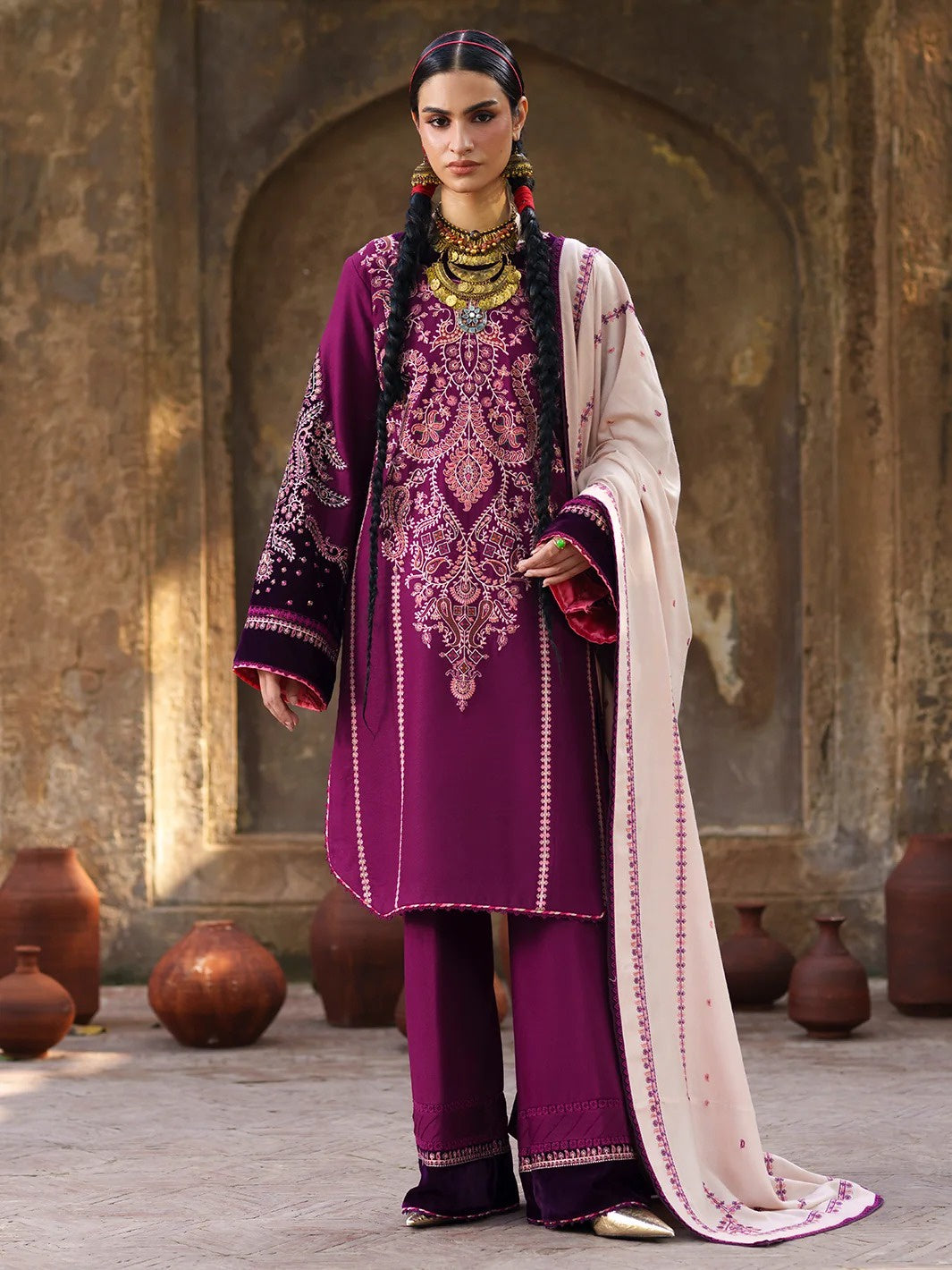 A woman models a traditional South Asian outfit consisting of a deep purple kurta with intricate embroidery, matching wide-leg trousers, and a light beige dupatta draped over her shoulder, set against a rustic stone archway background with terracotta pots.