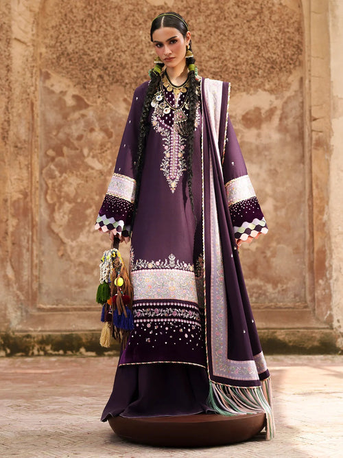 A woman models a traditional South Asian outfit consisting of a deep purple kurta with intricate silver and white embroidery, a matching purple dupatta with silver borders, and a brown embroidered tassel accessory, set against a textured stone wall background.