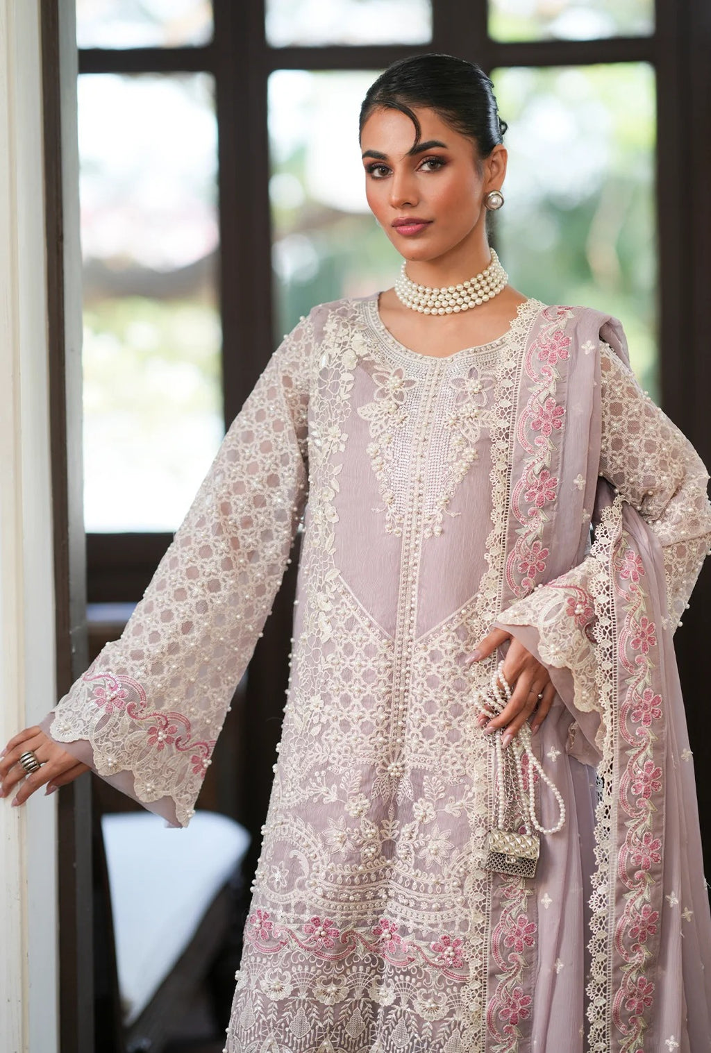A woman models a traditional three-piece salwar kameez suit in a soft lavender-gray color with intricate white floral embroidery and pink floral accents. The set includes a kurta, a matching jacket, and a sheer dupatta, styled with a pearl necklace and earrings.