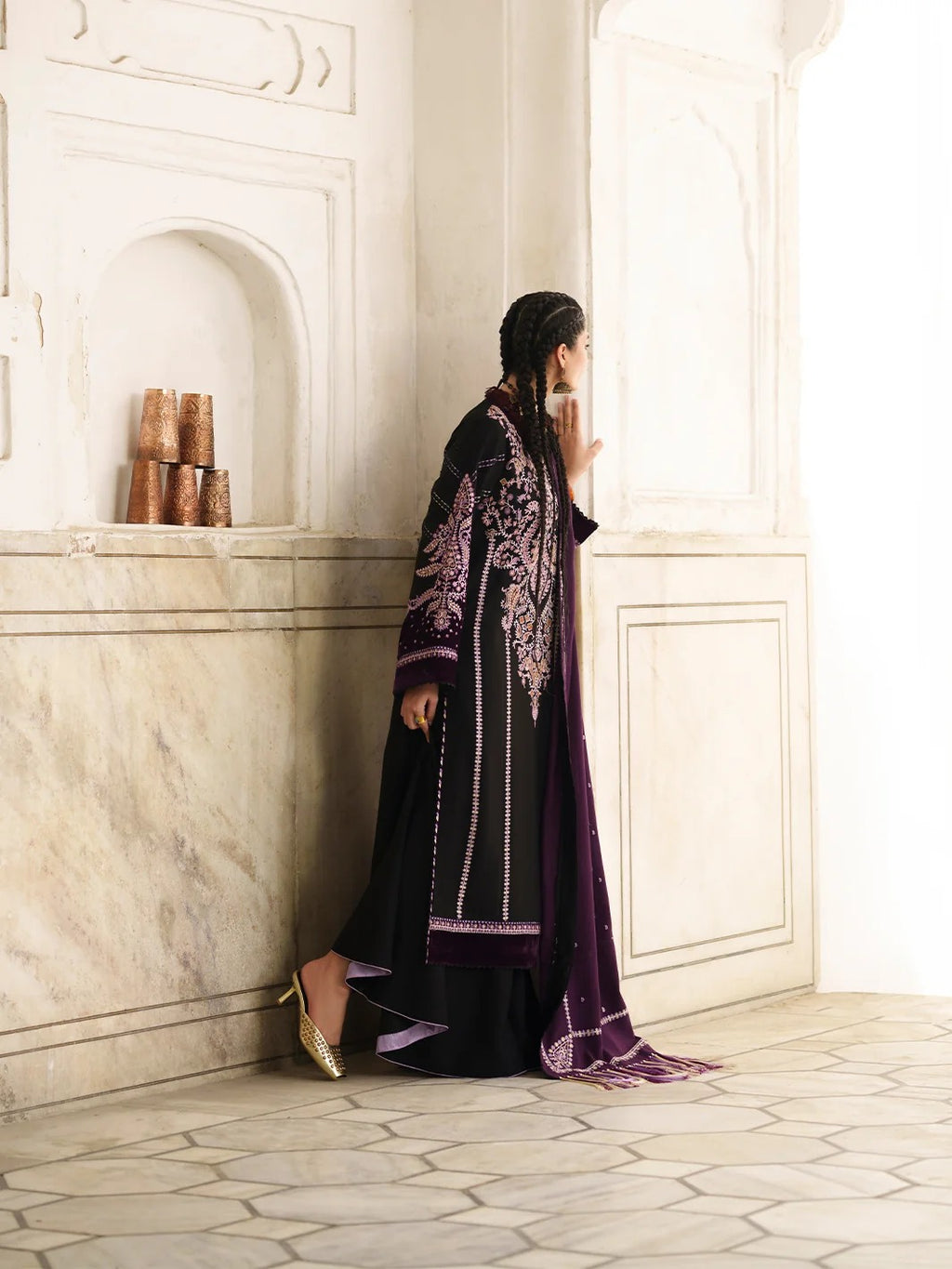 A woman wearing a black and purple embroidered traditional outfit with gold heels, standing in a light-colored architectural interior with marble walls and tiled floors.