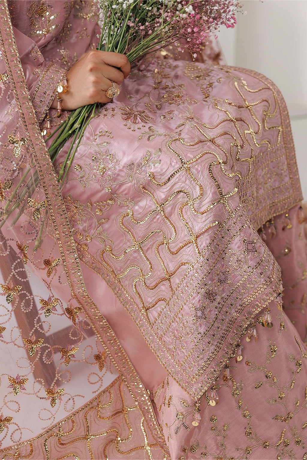 A close-up of a pink traditional embroidered garment with metallic gold embroidery and a matching sheer dupatta, held by a hand holding a bouquet of pink flowers.