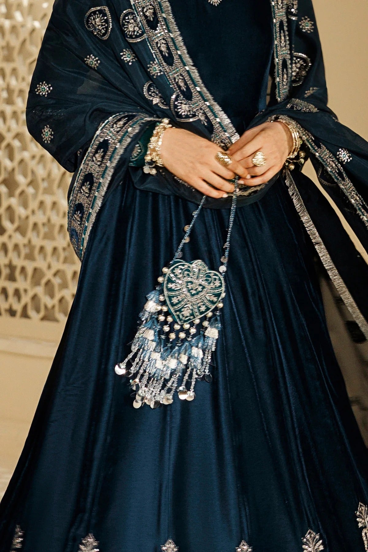 A close-up of a person wearing a dark navy blue traditional outfit with intricate silver embroidery, holding a matching embellished heart-shaped purse with dangling silver tassels.