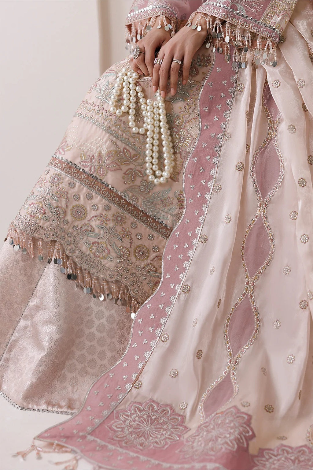 Close-up of a person wearing an ornate pink and beige traditional outfit with intricate embroidery, mirror work, and a pearl beaded necklace draped over the lapel.