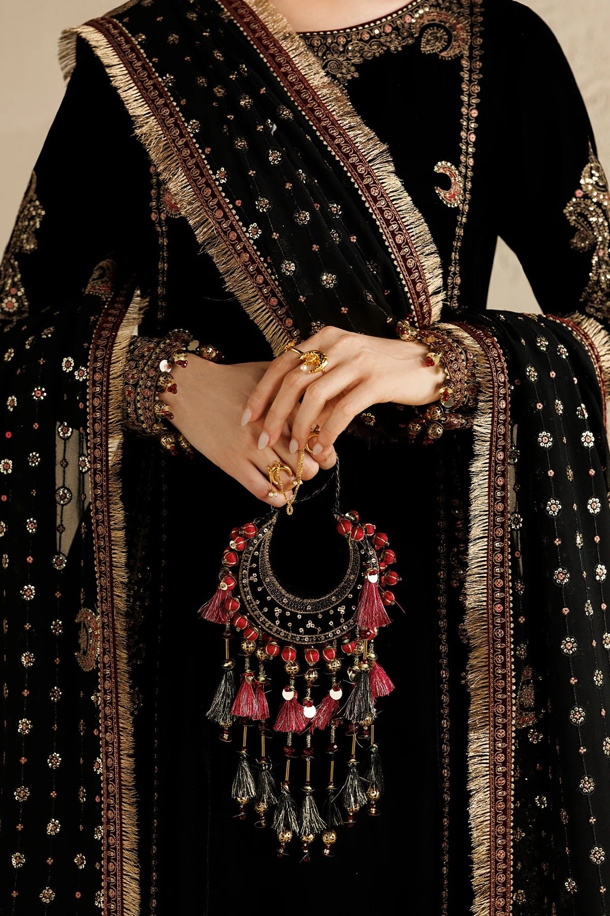 A close-up of a person wearing a black traditional outfit with ornate gold embroidery and a matching embroidered dupatta, holding a large gold-toned crescent pendant with red tassels and dangling charms.