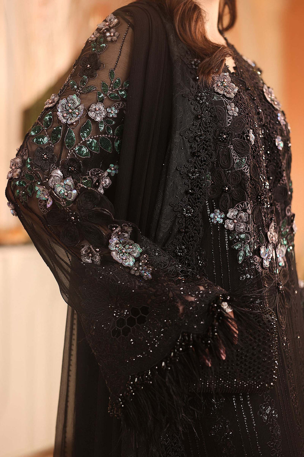 A close-up of a black sheer embroidered garment with floral beadwork and sequins, worn by a model in an indoor setting.