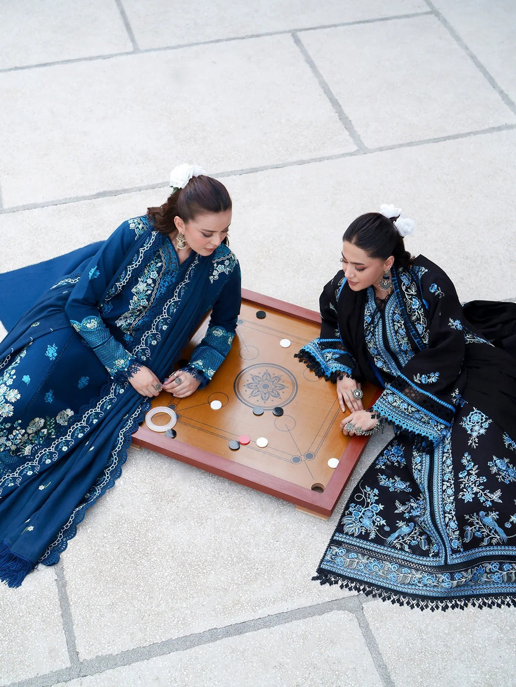 Two women in traditional embroidered clothing sit on a stone floor playing a board game. One wears a deep blue gown with white floral embroidery, and the other wears a black gown with blue embroidery.