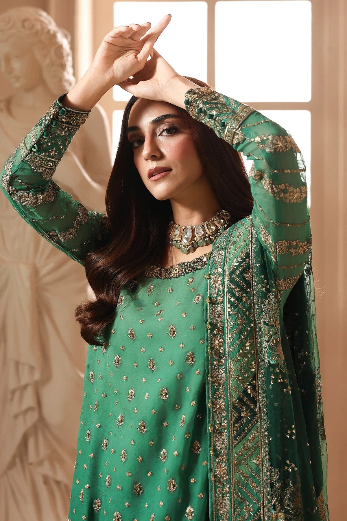 A woman modeling a green traditional embroidered outfit with metallic gold detailing, a matching dupatta, and a statement necklace in a warm indoor setting with a classical statue in the background.