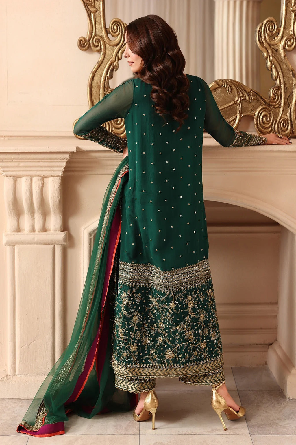 A woman modeling a dark green ethnic salwar kameez suit with gold embroidery and a matching dupatta, posed in front of an ornate cream-colored fireplace.