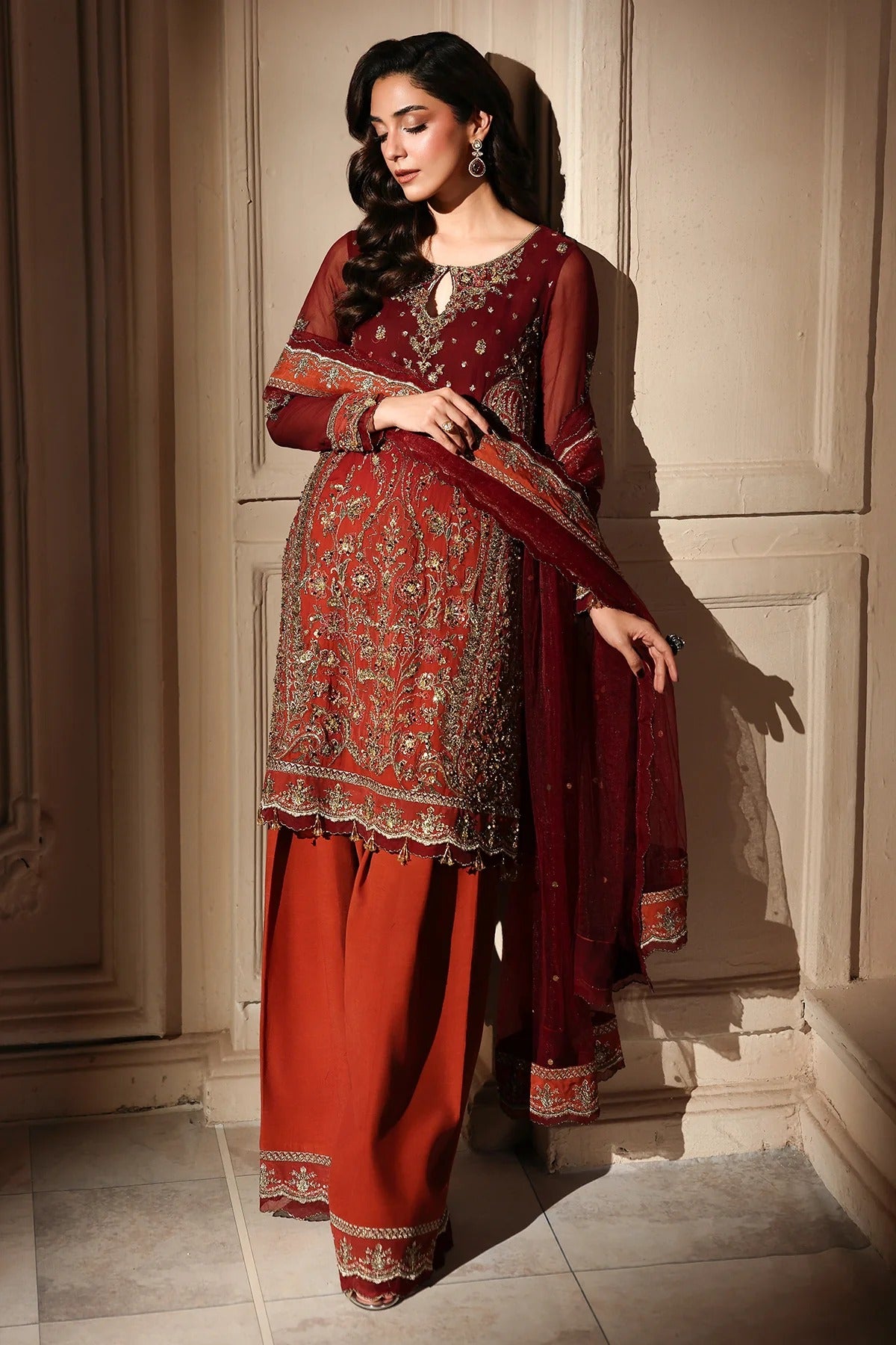 A woman models a traditional Indian salwar kameez suit set in a warm, elegant interior. The outfit features a deep maroon embroidered kameez jacket with gold and silver detailing, paired with rust-orange wide-leg trousers and a matching maroon dupatta.
