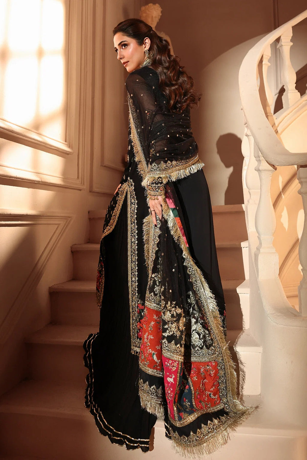 A woman modeling a black traditional outfit with ornate gold embroidery and red patterned panels, standing on indoor stairs near a white railing.