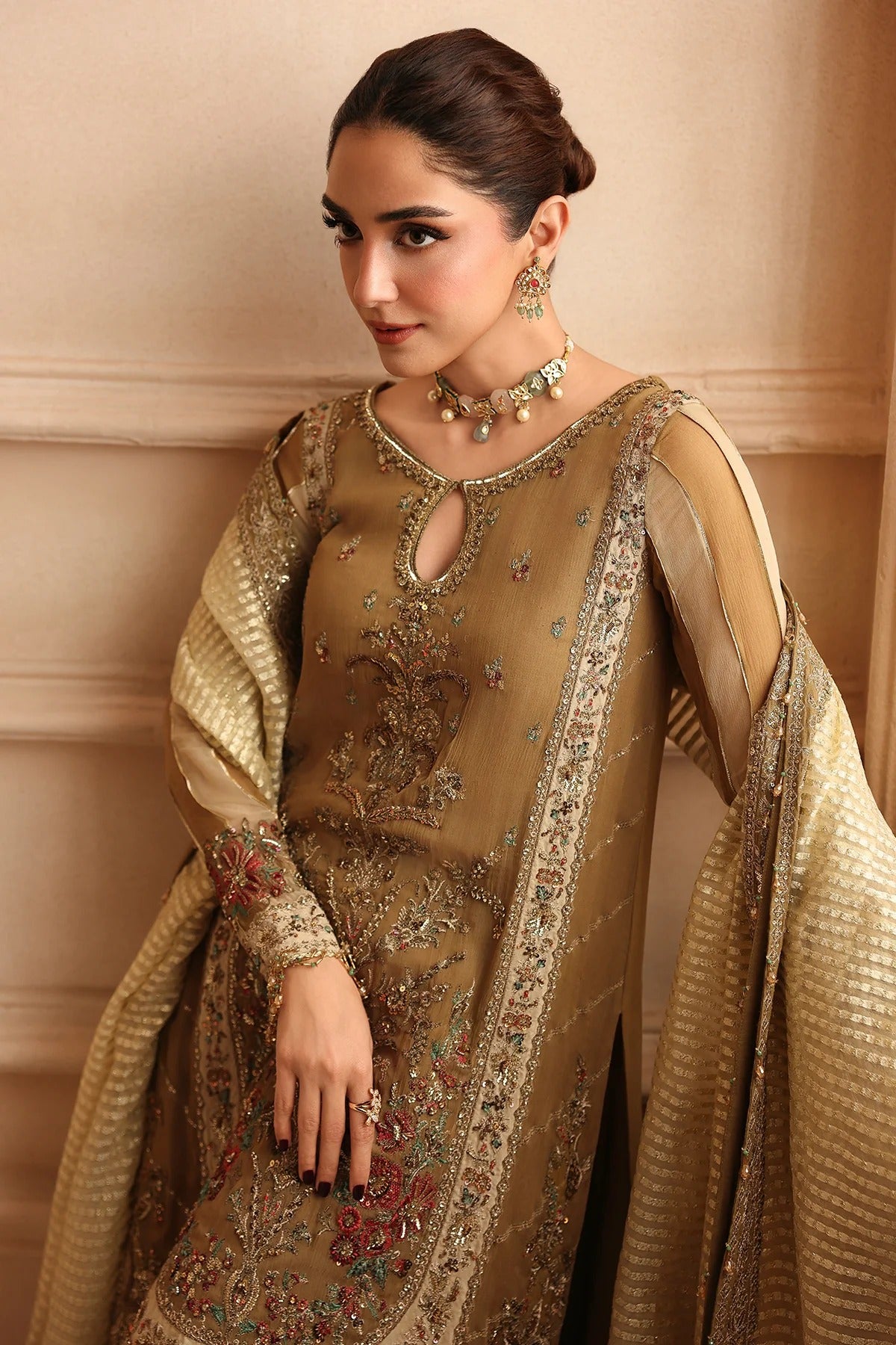A woman wearing an ornate beige and gold traditional embroidered outfit with a matching sheer dupatta, photographed against a warm beige architectural background.