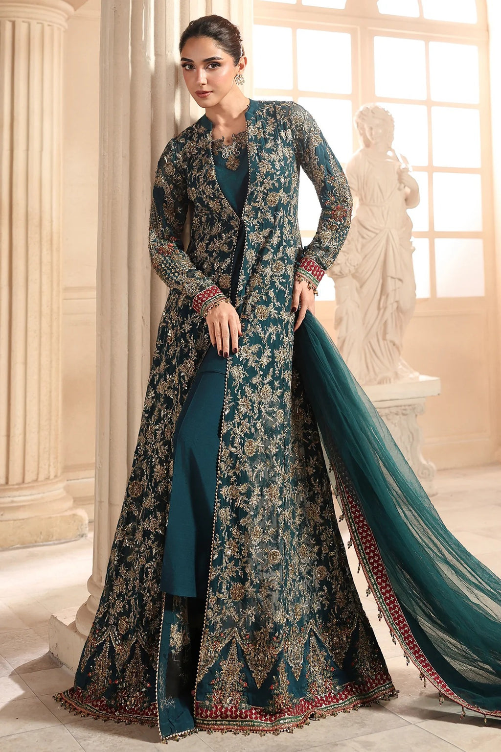 A woman models a traditional South Asian outfit consisting of a dark teal embroidered kurta, a matching long open-front jacket, and a sheer teal dupatta, set against a classical interior with columns and a statue.