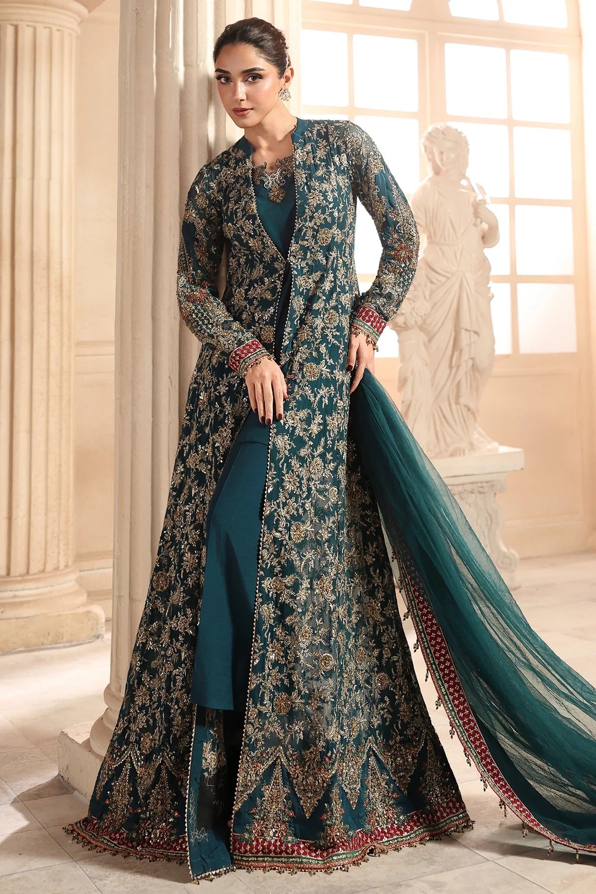 A woman models a traditional South Asian outfit consisting of a dark teal embroidered kurta, a matching long open-front jacket, and a sheer teal dupatta, set against a classical interior with columns and a statue.