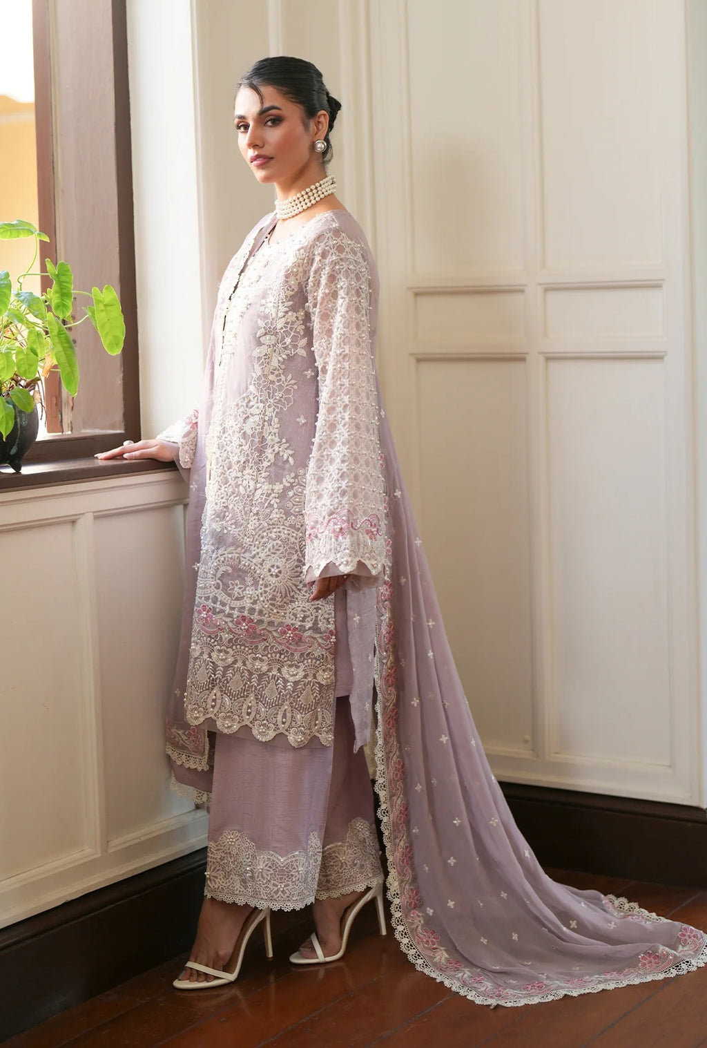 A woman models a traditional Indian outfit consisting of a lavender kurta with intricate white and pink floral embroidery, matching lavender pants, and a long sheer lavender dupatta with small gold motifs, set against an indoor background with a window and paneled wall.