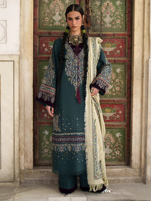 A woman models a traditional South Asian outfit consisting of a dark teal embroidered kurta, matching trousers, and a cream dupatta with gold embroidery, standing in front of an ornate green and red wooden door.