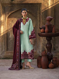 A woman models a traditional South Asian salwar kameez suit in a rustic courtyard setting. The outfit features a mint green kurta with deep red and gold floral embroidery, paired with a deep maroon dupatta that has matching embroidered borders.