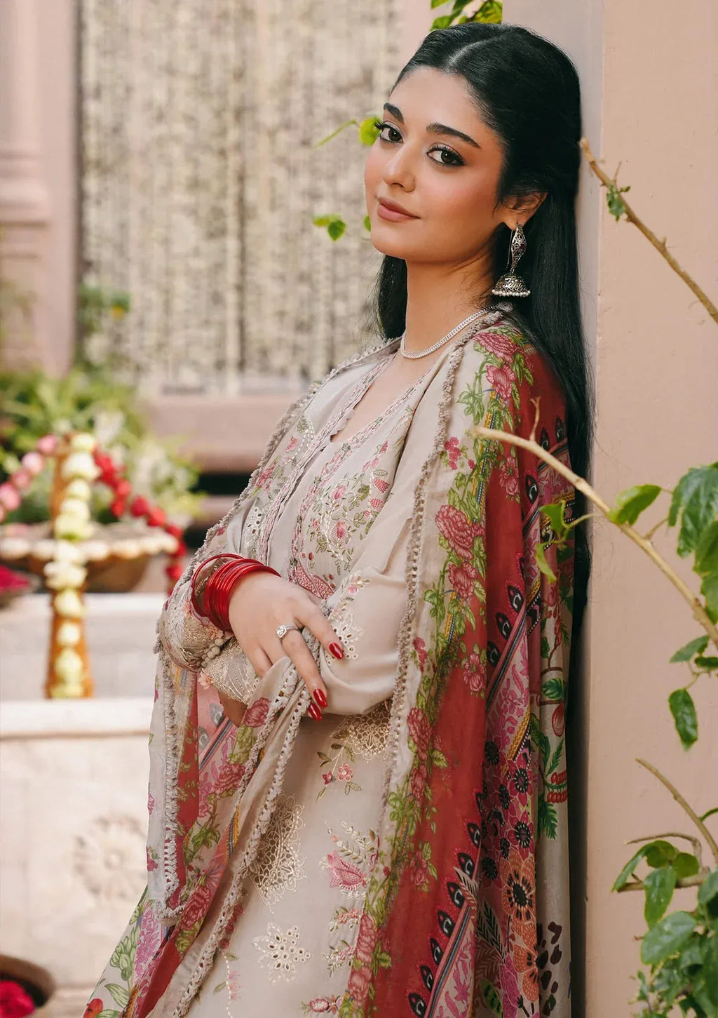 A woman wearing a traditional South Asian salwar kameez outfit in beige with pink and green floral prints, standing in an outdoor courtyard setting.