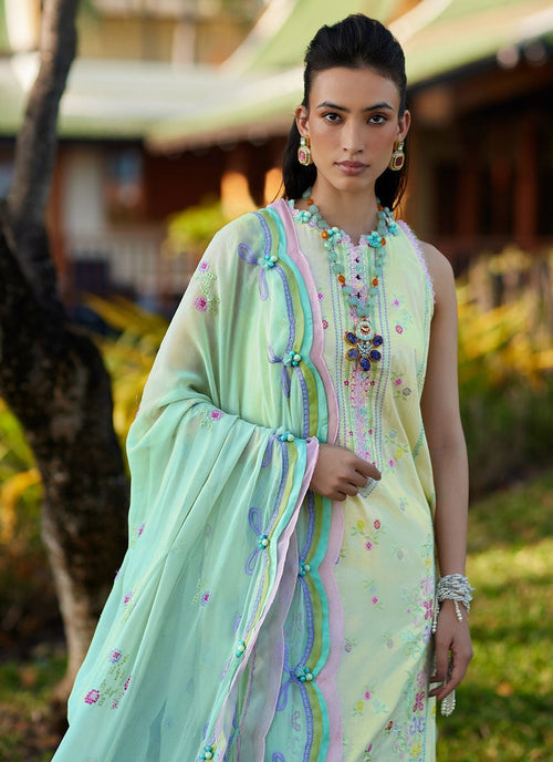 A woman modeling a traditional light green ethnic outfit with pastel pink, blue, and yellow embroidered borders and a matching sheer dupatta, photographed outdoors in a garden setting.