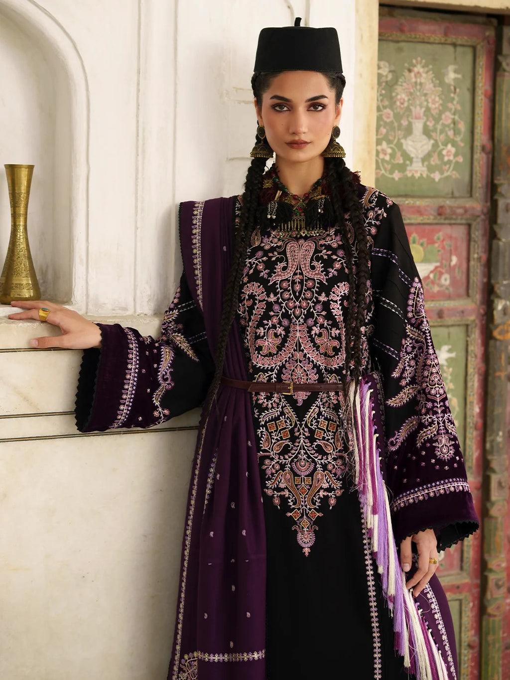 A woman models a traditional embroidered outfit with a black base, deep purple sleeves and dupatta, and intricate pink and gold embroidery, set against an ornate architectural background.