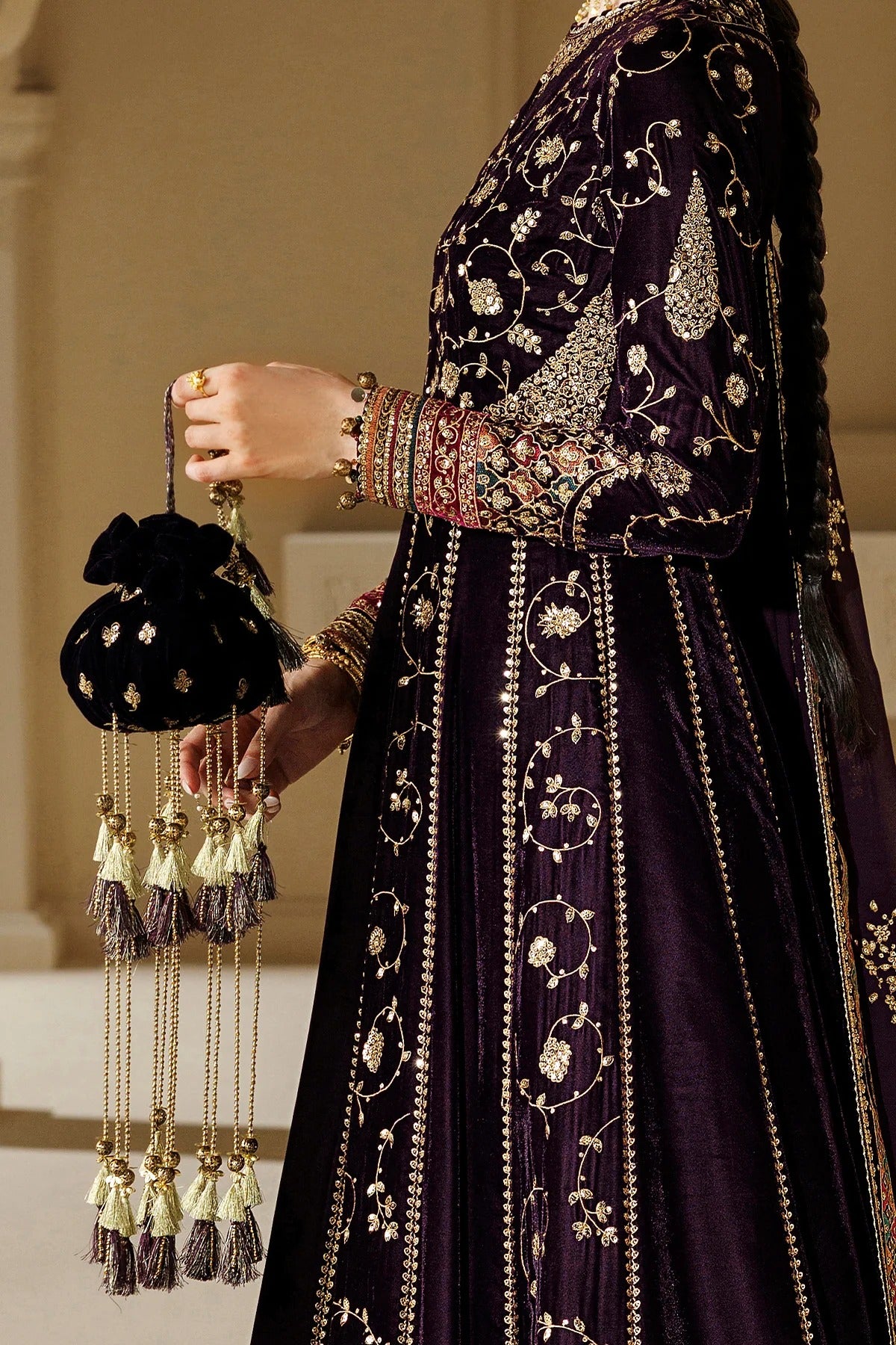 A close-up of a person wearing an ornate deep purple embroidered traditional outfit with gold detailing, holding a small black embroidered pouch with tassel charms.
