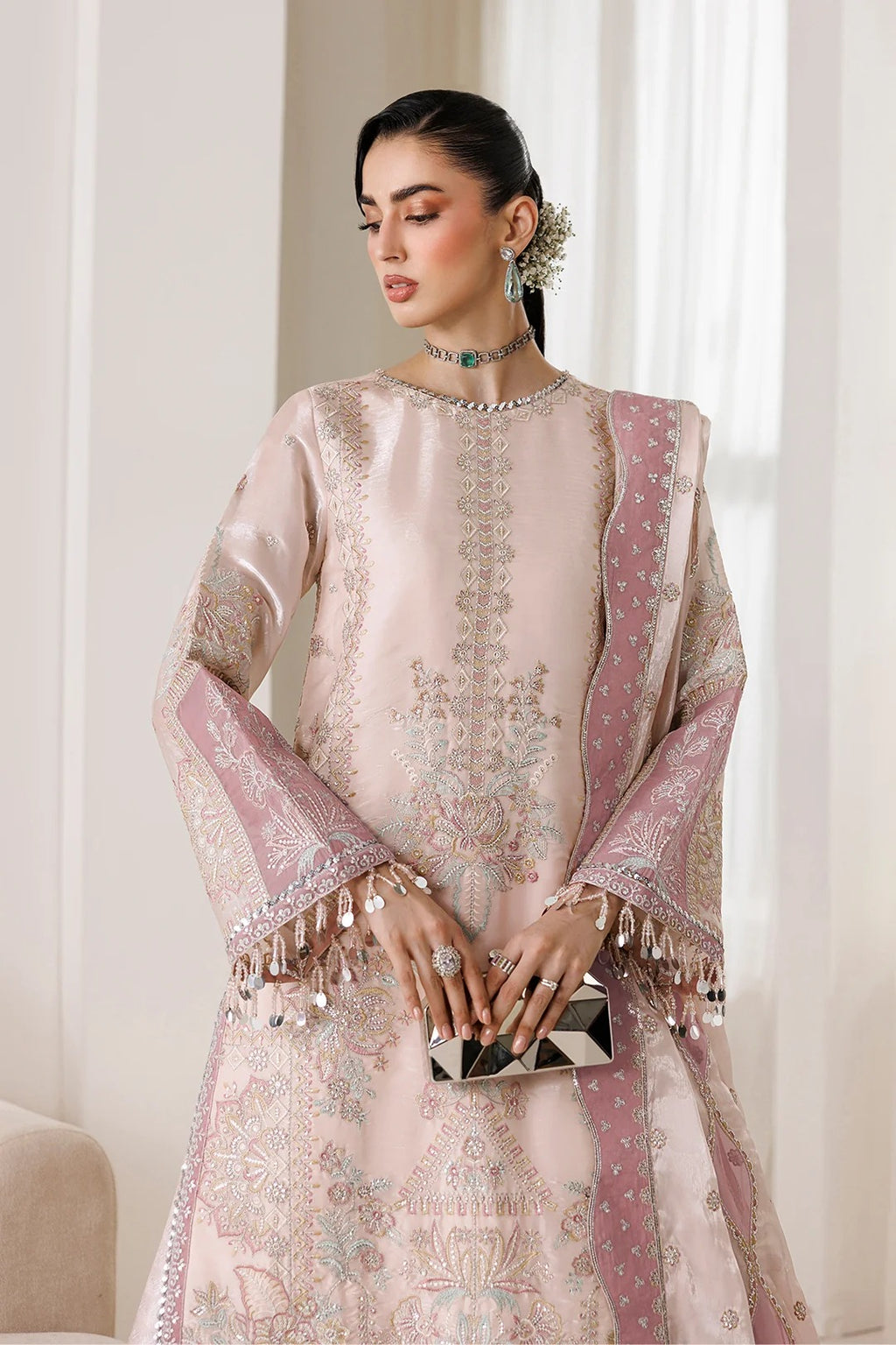 A woman models a traditional light pink embroidered outfit with matching dupatta, featuring intricate metallic embroidery, silver fringe, and a small black-and-white clutch, set against a soft beige indoor background.