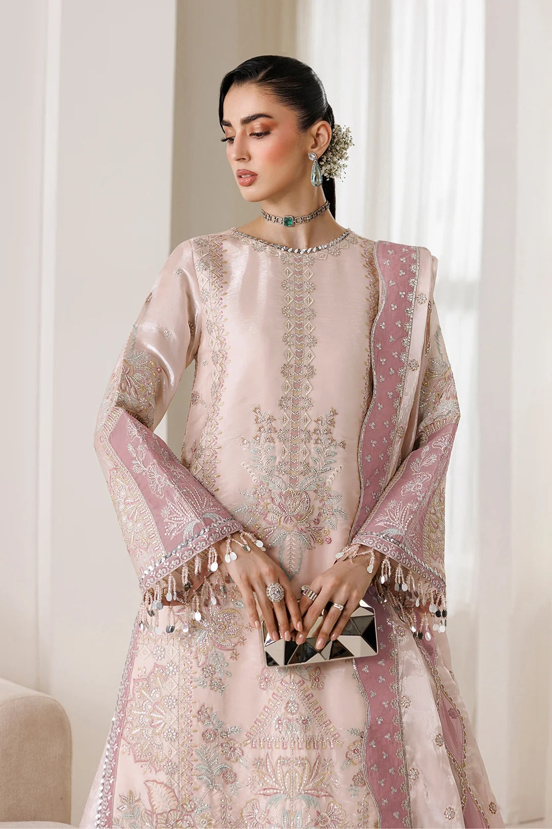 A woman models a traditional light pink embroidered outfit with matching dupatta, featuring intricate metallic embroidery, silver fringe, and a small black-and-white clutch, set against a soft beige indoor background.