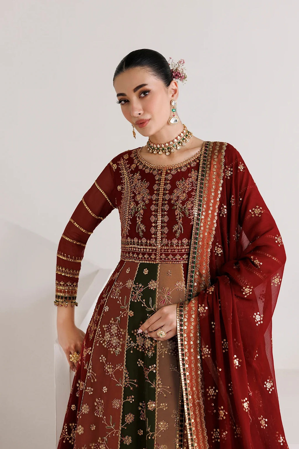A woman models a traditional Indian salwar kameez outfit in deep maroon and dark green with extensive gold embroidery, paired with a matching sheer dupatta, set against a plain light gray background.