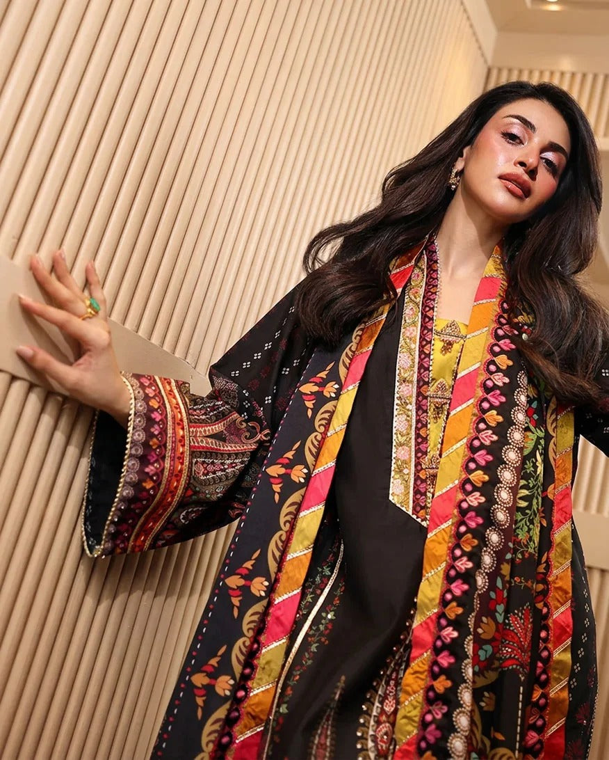 A woman models a traditional South Asian outfit featuring a black kurta with ornate multicolor embroidery, paired with a matching patterned dupatta and a black coat draped over her shoulder, set against a modern slatted wood wall.