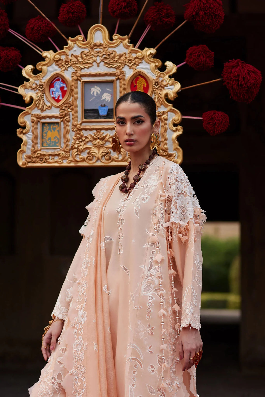 Woman in a peach traditional outfit with intricate embroidery, standing in front of a decorative backdrop.
