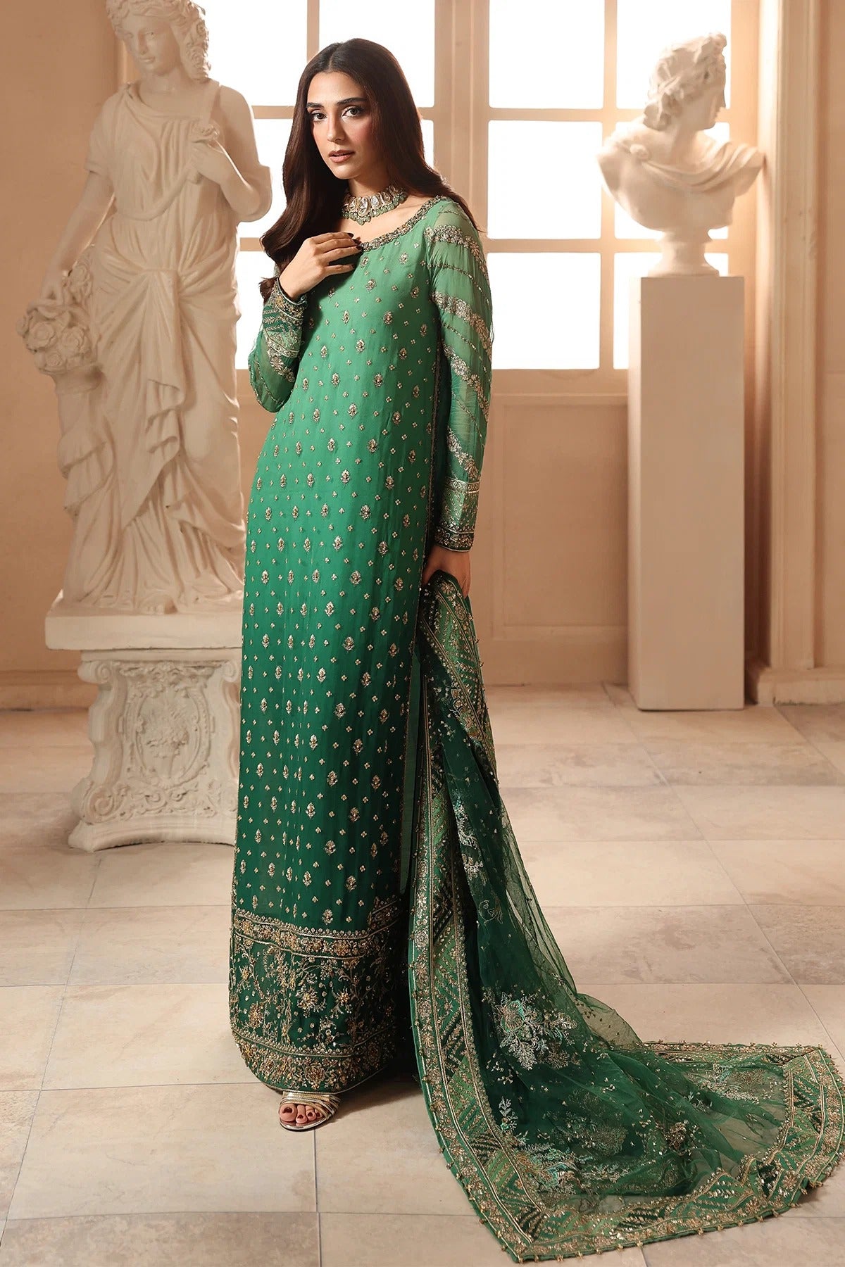 A woman models a traditional green salwar kameez suit with gold embroidery and a matching dupatta in an elegant indoor setting with classical statues.