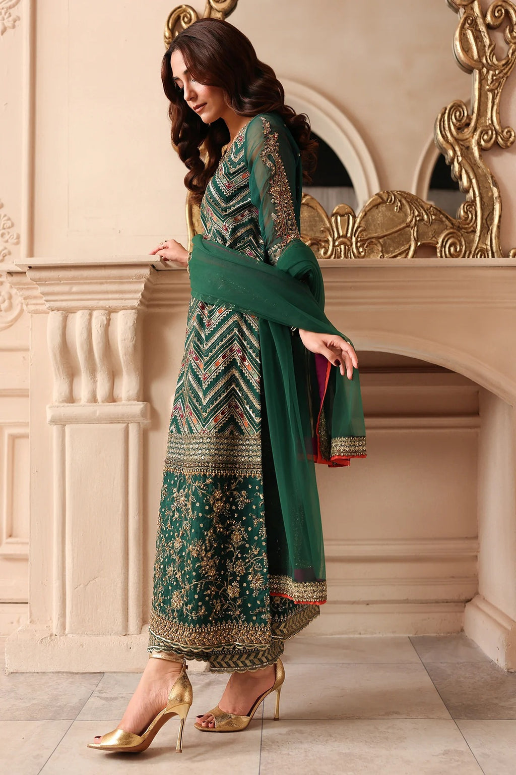 A woman models a traditional green salwar kameez suit with intricate gold and silver embroidery, paired with a matching green dupatta and gold high heels, set against an ornate beige stone wall and mirror.