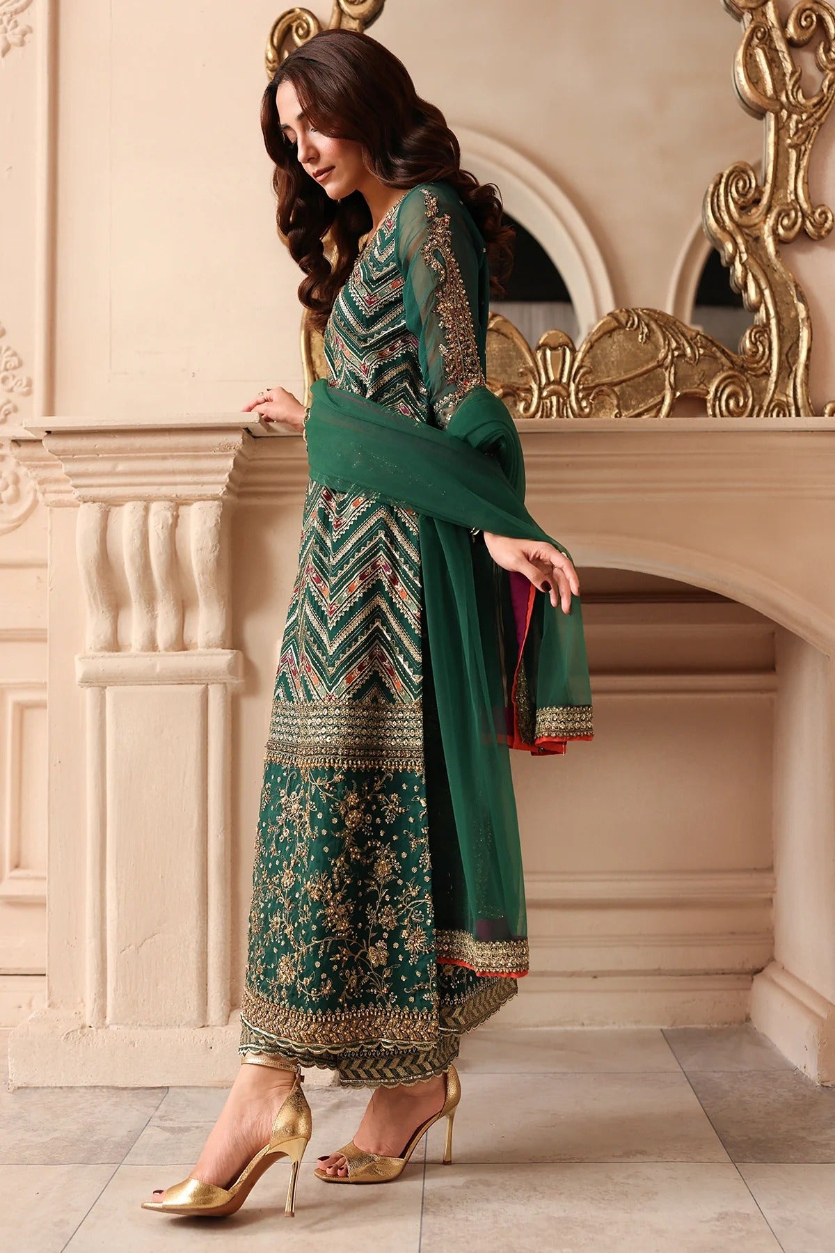 A woman models a traditional green salwar kameez suit with intricate gold and silver embroidery, paired with a matching green dupatta and gold high heels, set against an ornate beige stone wall and mirror.