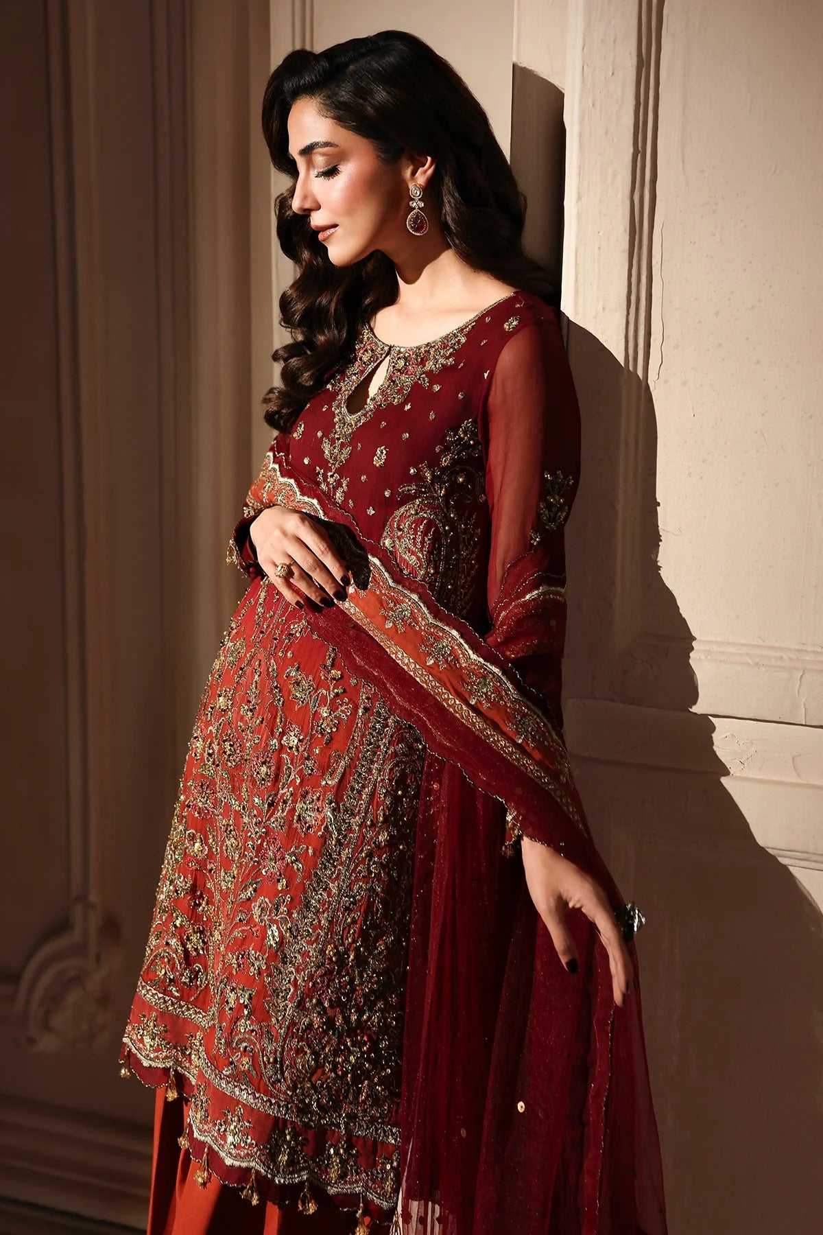 A woman models a traditional red and gold embroidered ethnic outfit with a matching dupatta, posed against a beige wall with dramatic side lighting.