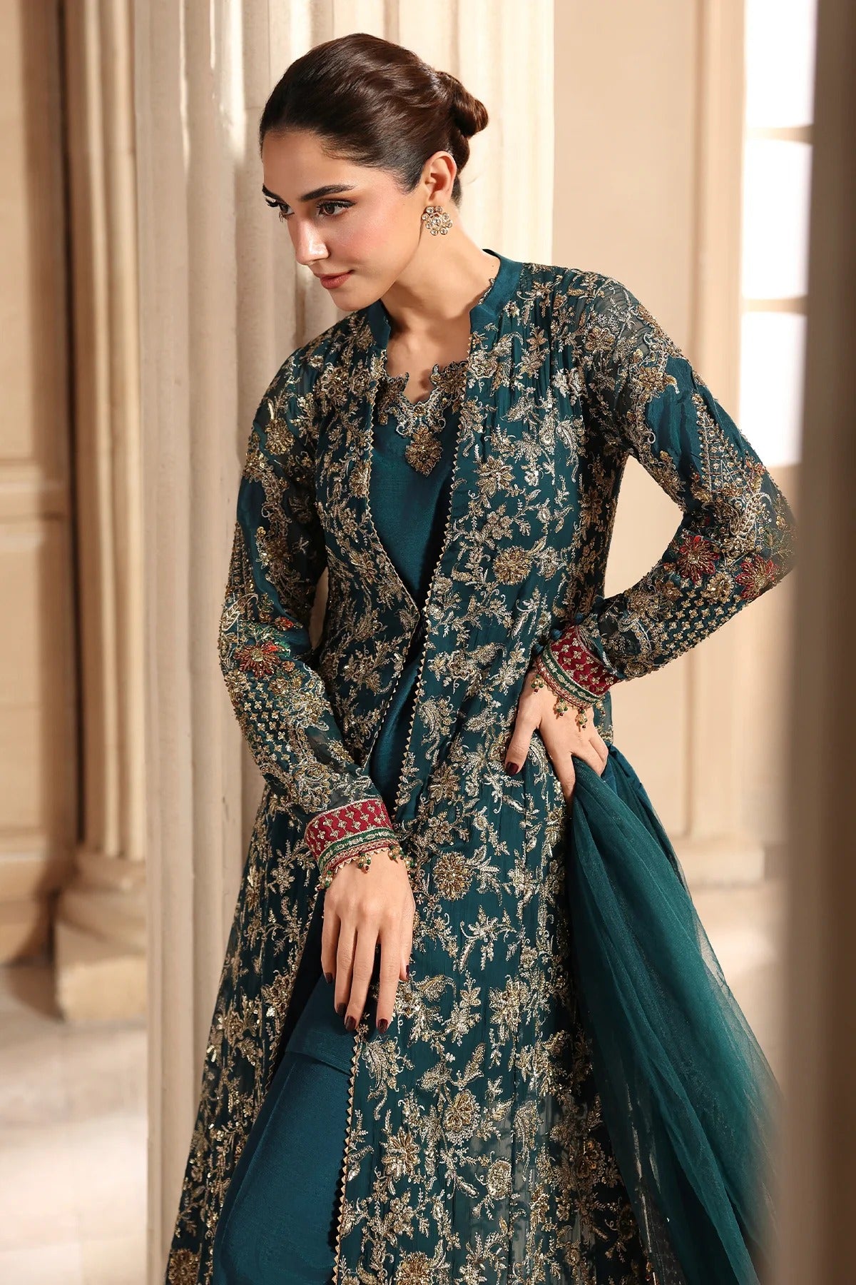 A woman models a traditional South Asian outfit featuring a dark teal embroidered kurta with a matching dupatta, set against a warm beige architectural background.