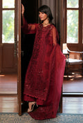 A woman models a deep maroon red embroidered traditional outfit with sheer sleeves and a matching dupatta, standing in a doorway of a wooden house.